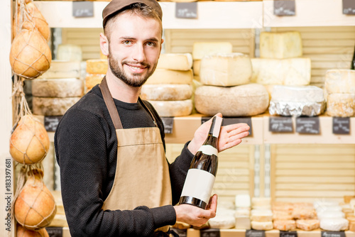 Portrait of a young sommelier in uniform standing with wine bottle in front of the store showcase full of different cheeses photo