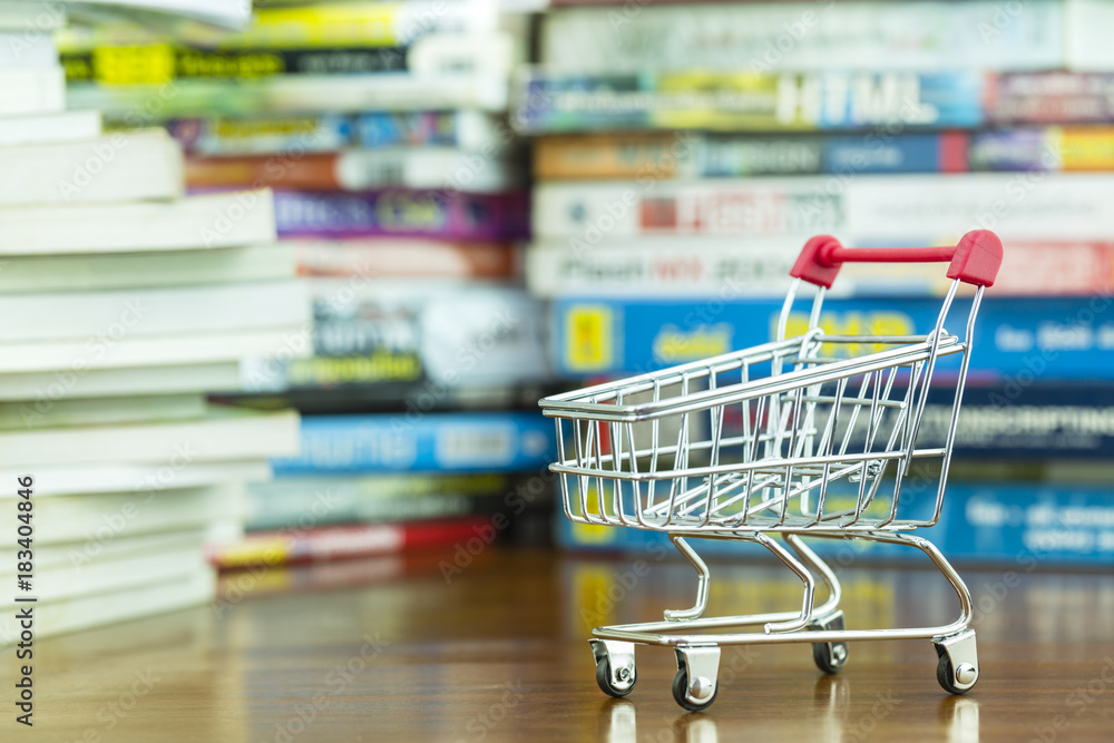 Mini shopping cart on wooden table with blurred pile of book as background, concept of shopping online knowledge to advance Mini shopping cart on wooden table with blurred pile of book as background, concept of shopping online knowledge to advance