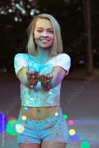 Beautiful smiling woman covered with dry Holi powder posing with colorful blurry lights of garland