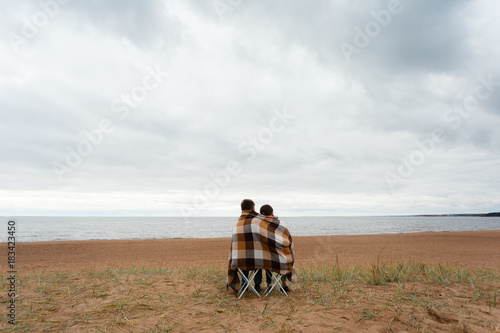 Rear view of a couple sitting on the shore of natural beach wrapped up sharing a blanket on a cold winter vacation, outdoors space. Boyfriend and girlfriend travel, serenity and contemplation.