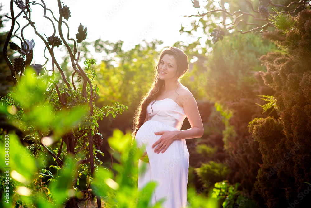 Naklejka premium A happy pregnant girl in a white dress is walking in the park.