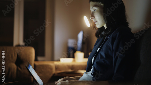 Young concentrated woman working at night using laptop computer and typing message