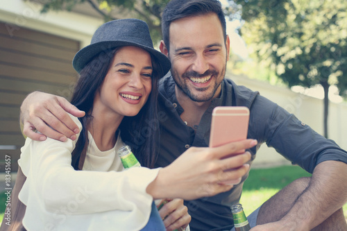 Happy young couple sitting in city park drinking beer and take a selfie.