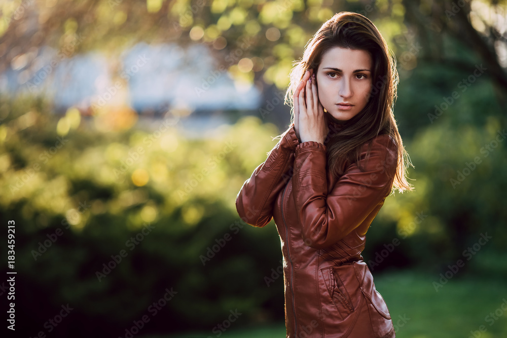 Young romantic woman in brown leather jacket over background early spring portrait. Pretty girl posing in park. Fashionable long hair model in elegant clothes. Copy space