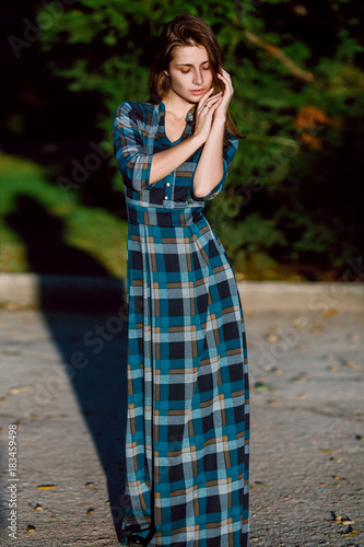 A woman in long checkered dress. Romantic girl in the spring park. A woman walks in the park in a casual dress. Green field on background. Vertical orientation