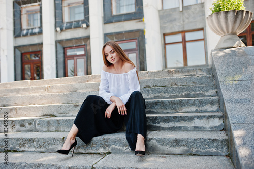 Good-looking young woman in white blouse, wide black pants and black classic high heels sitting on stairs and posing.