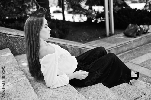 Good-looking young woman in white blouse, wide black pants and black classic high heels sitting on stairs and posing. Black and white photo.
