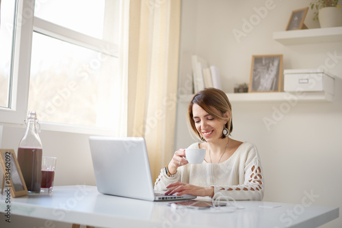 Woman drinking coffee and using laptop for work while sitting at home office