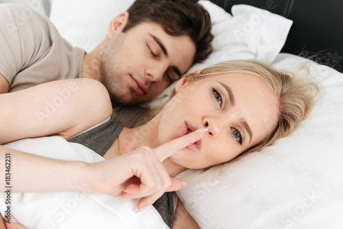 Young beautiful blonde lady showing silence gesture while lying in bed with her man