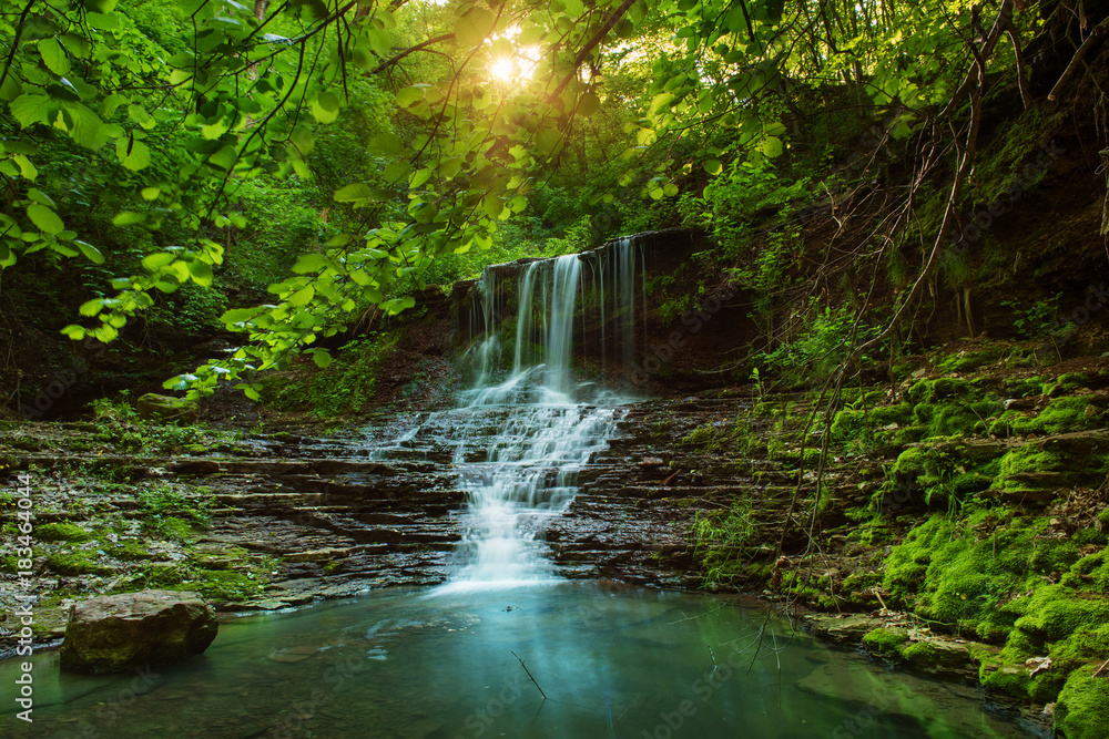 Beautiful mountain rainforest waterfall with fast flowing water and rocks, long exposure. Natural seasonal travel outdoor background with sun shihing
