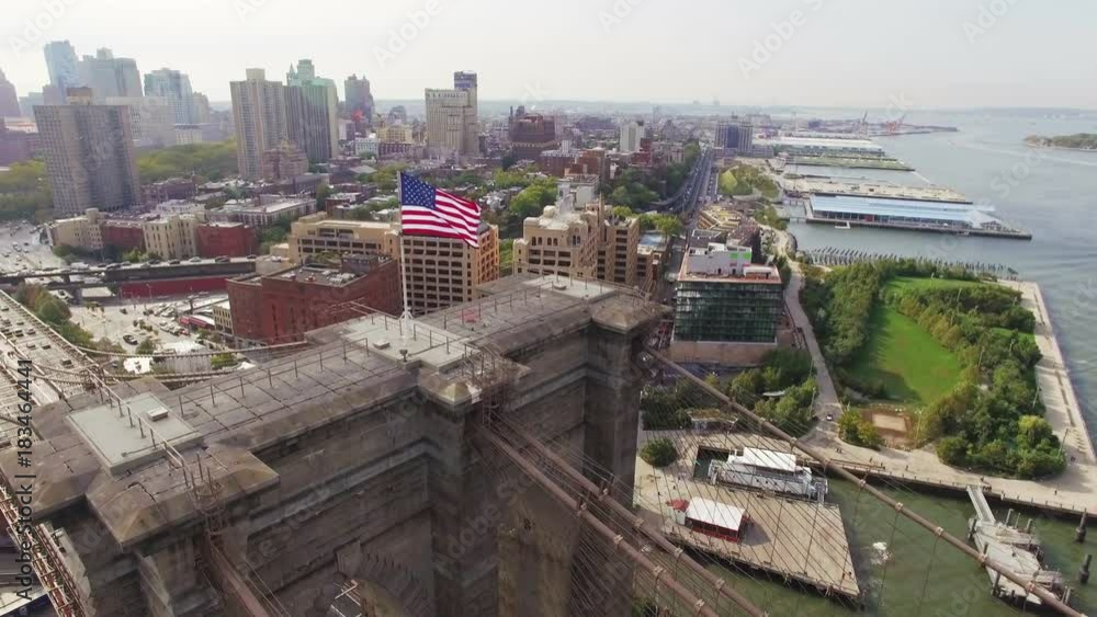 Brooklyn bridge, aerial shot. Camera moving around a flapping American flag to Brooklyn across the East river, Diamond Reef view. Cars moving across the bridge. Brooklyn bridge, aerial shot. Camera moving around a flapping American flag to Brooklyn across the East river, Diamond Reef view. Cars moving across the bridge.