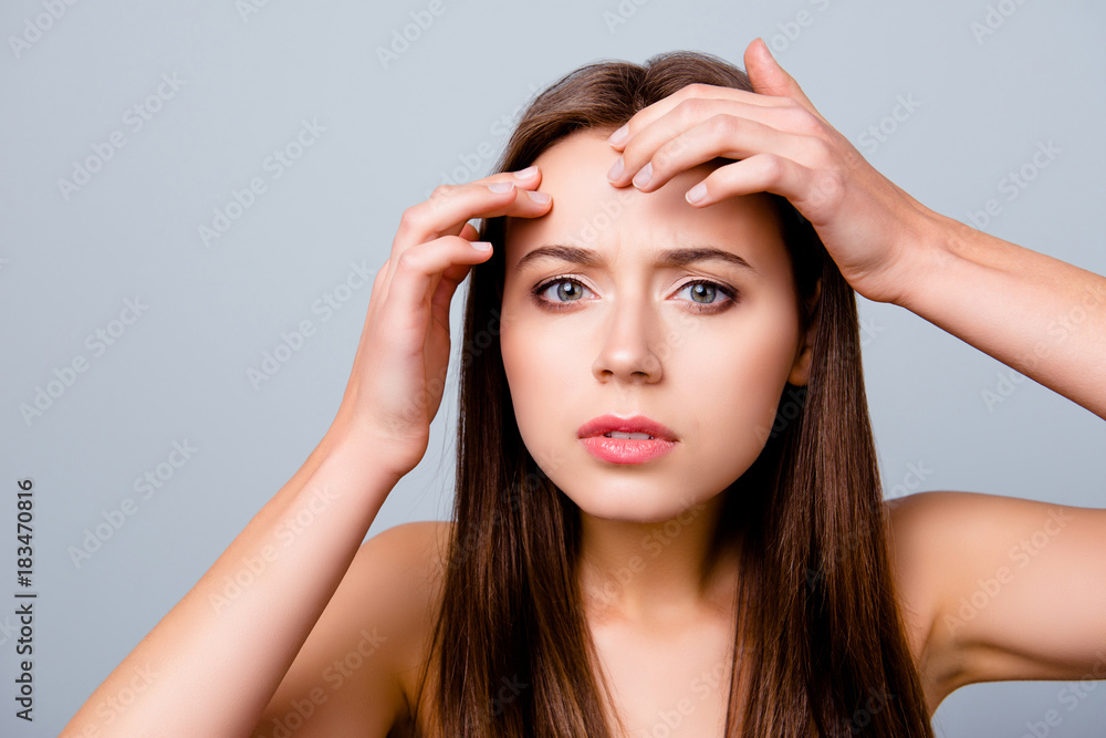 Fototapeta premium Close up portrait of frustrated sad upset beautiful young woman is squeezing out pimples on her forehead, isolated on grey background