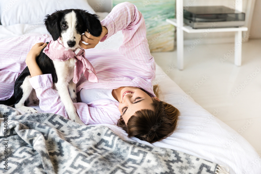 young woman in pajamas lying on bed together with black and white puppy young woman in pajamas lying on bed together with black and white puppy