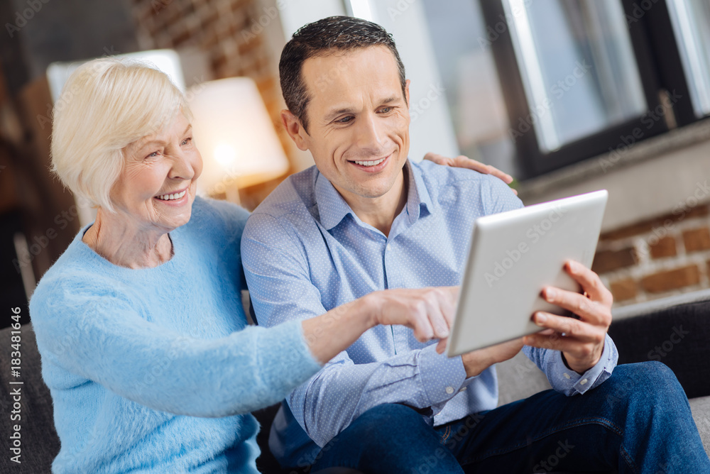 Becoming technology-savvy. Petite senior woman sitting on the couch next to her son and tapping the tablet while her son teaching her how to use it Becoming technology-savvy. Petite senior woman sitting on the couch next to her son and tapping the tablet while her son teaching her how to use it