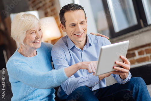 Becoming technology-savvy. Petite senior woman sitting on the couch next to her son and tapping the tablet while her son teaching her how to use it