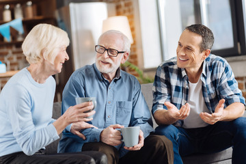 Enjoying themselves. Cheerful happy family consisting of elderly couple and their adult son sitting on the sofa, drinking tea and communicating with each other