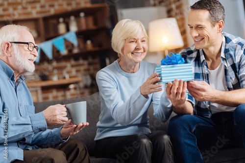 Gift with love. Cheerful young man giving a birthday present to his happy elderly mother while his father sitting next to them and drinking coffee