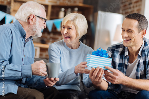 Happy birthday. Upbeat young man sitting on the sofa next to his elderly parents and giving a birthday present to his mother while she sharing happiness with her husband
