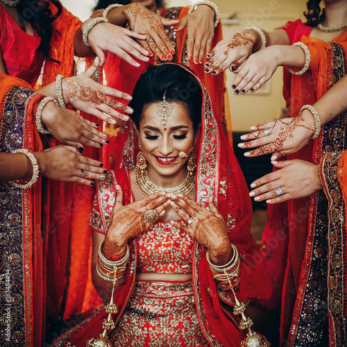 Stunning Indian bride dressed in Hindu traditional wedding clothes lehenga embroidered with gold and a veil sits on the chair while bridesmaids hold their hands with henna tattoos around her