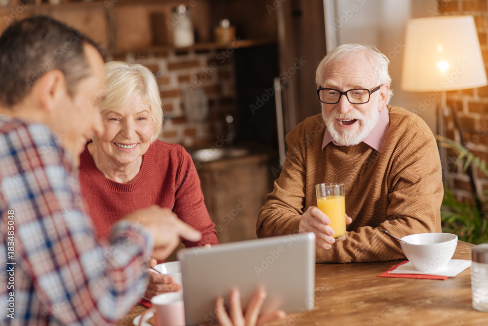 Gripping content. Cheerful young man using a tablet to show a video to his elderly parents while they all having breakfast in the kitchen Gripping content. Cheerful young man using a tablet to show a video to his elderly parents while they all having breakfast in the kitchen