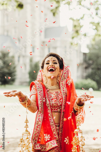 Stunning Indian bride dressed in Hindu traditional wedding clothes lehenga embroidered with gold and a veil smiles tender posing outside with golden accessories under the rain of petals