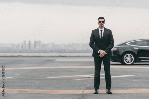 serious bodyguard standing with sunglasses and security earpiece on helipad and looking at camera photo