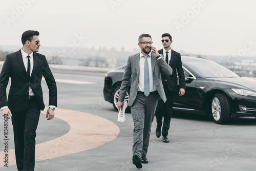 businessman walking with bodyguards on helipad and talking by smartphone photo