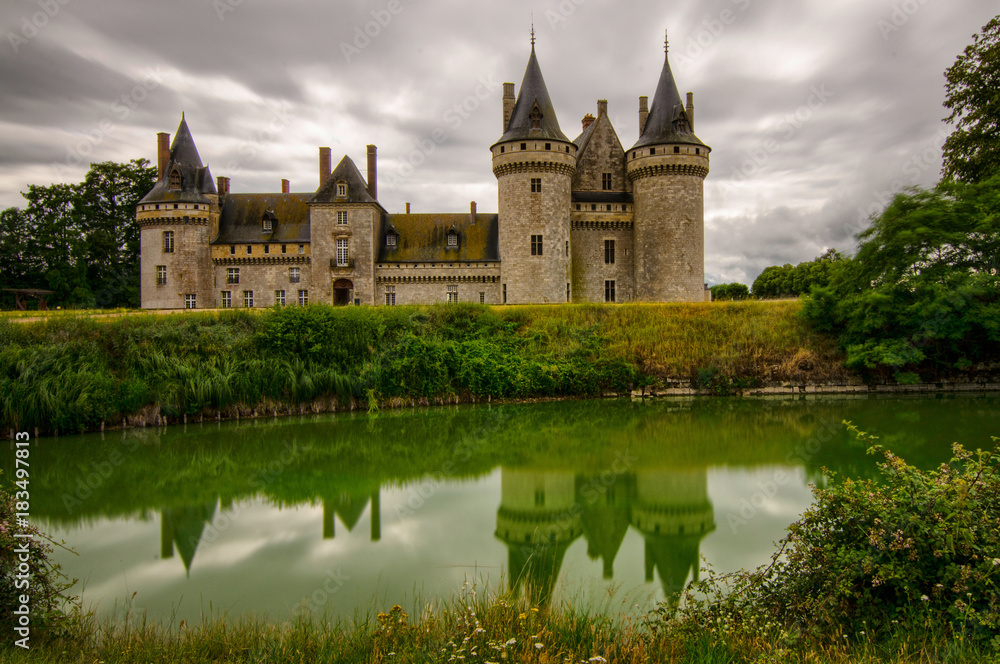 Castle of Sully-sur-Loire, Loire region, France. Snap of 30 June 2017 17:38. Shooting from the park towards the castle entrance. The façade is mirrored in the water of the moat. Sky with stormy clouds