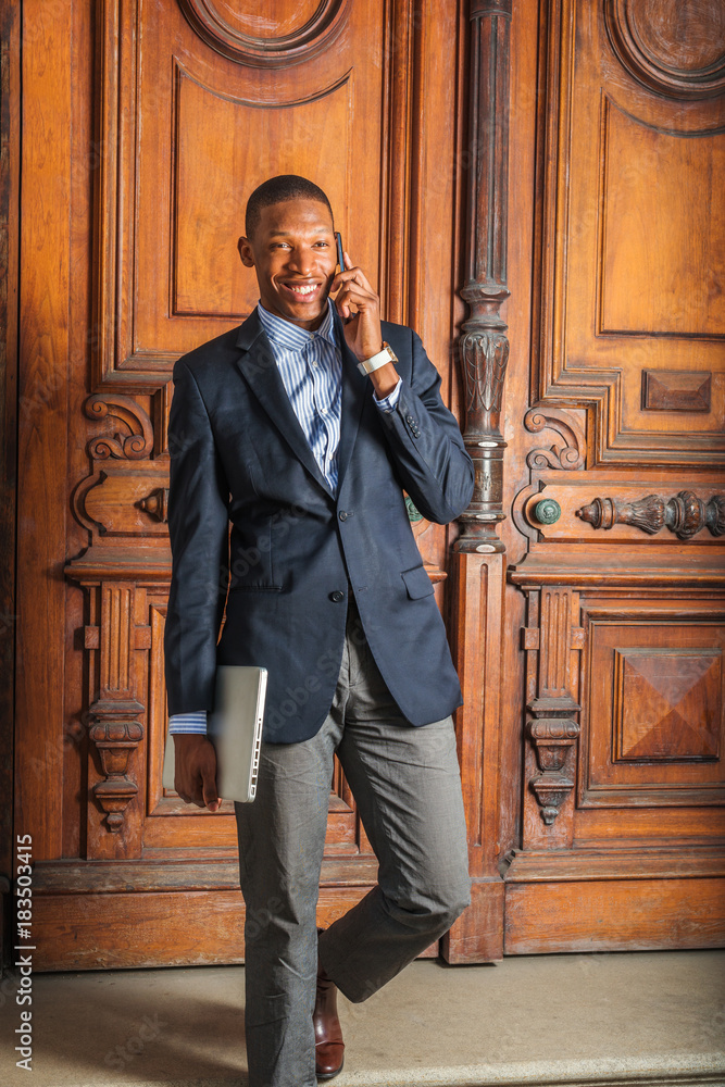 agenda talent Buzz Wearing black blazer, gray pants, brown leather shoes, hand carrying laptop  computer, a young African American businessman walking down stairs from  vintage style office door way, talking on phone. Stock Photo 