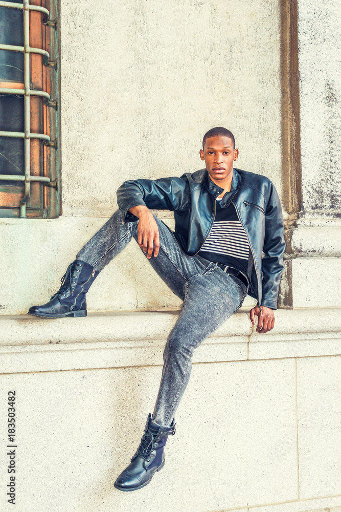 Man Urban Casual Fashion. Wearing black leather jacket, striped undershirt,  jeans, leather boots, a young African American guy sitting against wall on  street in New York, relaxing, waiting for you.. Stock Photo