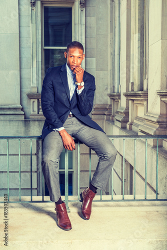 Dressing in black blazer, gray pants, brown leather shoes, wristwatch,  African American businessman sitting on railing in vintage style office  building, fist touching chin, thinking. . Stock Photo | Adobe Stock
