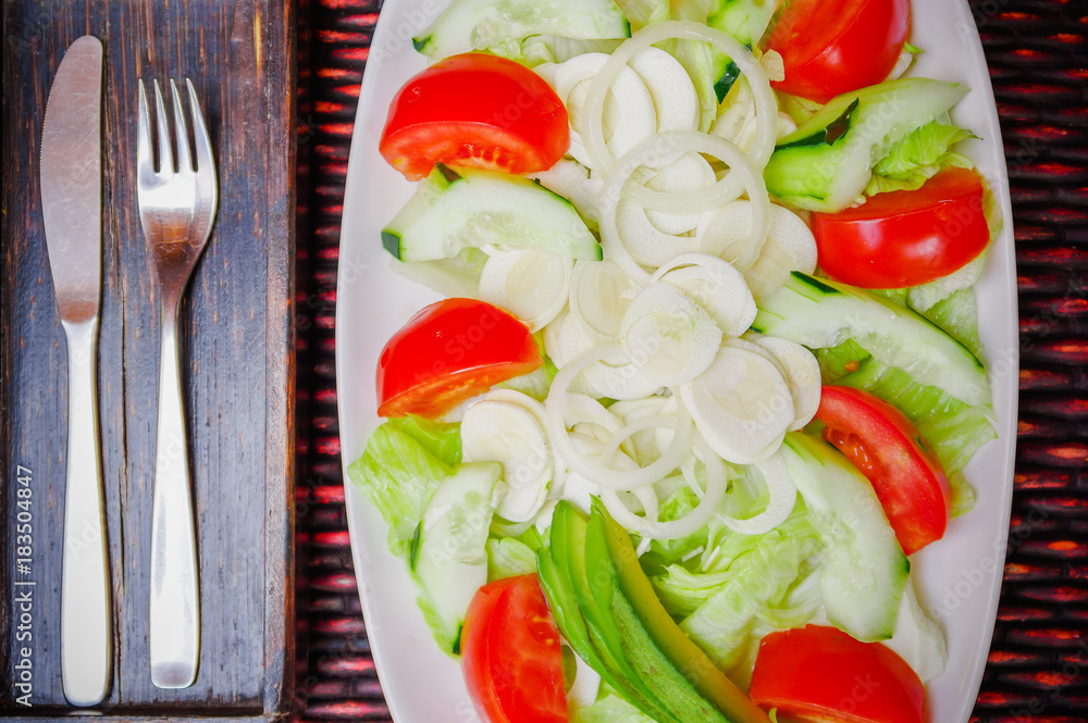 Above view of fresh salad with vegetables, tomato, onion, palmito, cucumbers, served on a white plate with a fork and knife in the table, on wooden table Above view of fresh salad with vegetables, tomato, onion, palmito, cucumbers, served on a white plate with a fork and knife in the table, on wooden table