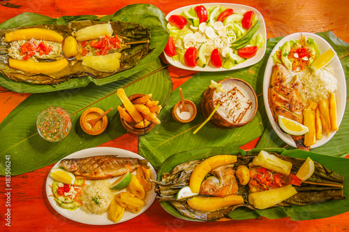 Above view of assorted food, grilled meat with potato, sweet tomatos, salad, pepper, fried plantain, fish in a leaf with yucca, fried fish, fried yucca, palmito salad, served in white plates over a photo