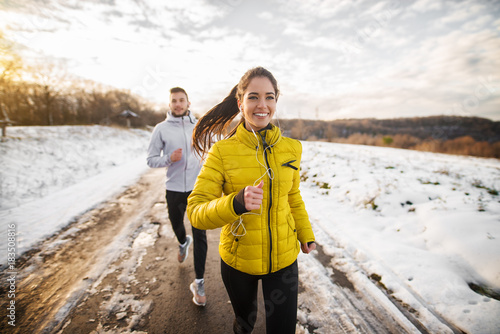 Beautiful happy active runner girl jogging with her personal handsome trainer on a snowy road in nature.