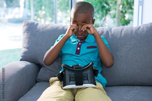 Boy rubbing eyes while sitting with VR headset on sofa photo