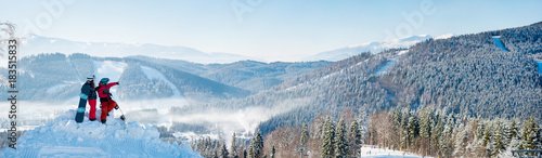 Winter panorama of the mountains landscape and forests in a white haze, two snowboarders resting on top of the mountain on a sunny winter day. Man pointing to the background copyspace snowboarding