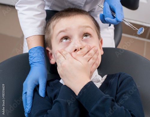 Close-up of boy frightened by dentists covers her mouth and looks for him. Doctor in gloves with one hand holds the instruments with the other hand supports the patient