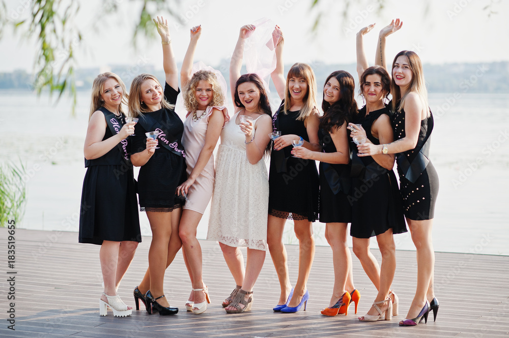 Fototapeta premium Group of 8 girls wear on black and 2 brides at hen party against sunny beach drinking champagne.