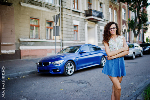 Curly stylish girl wear on blue jeans skirt, blouse and glasses posed near blue car at street of city.