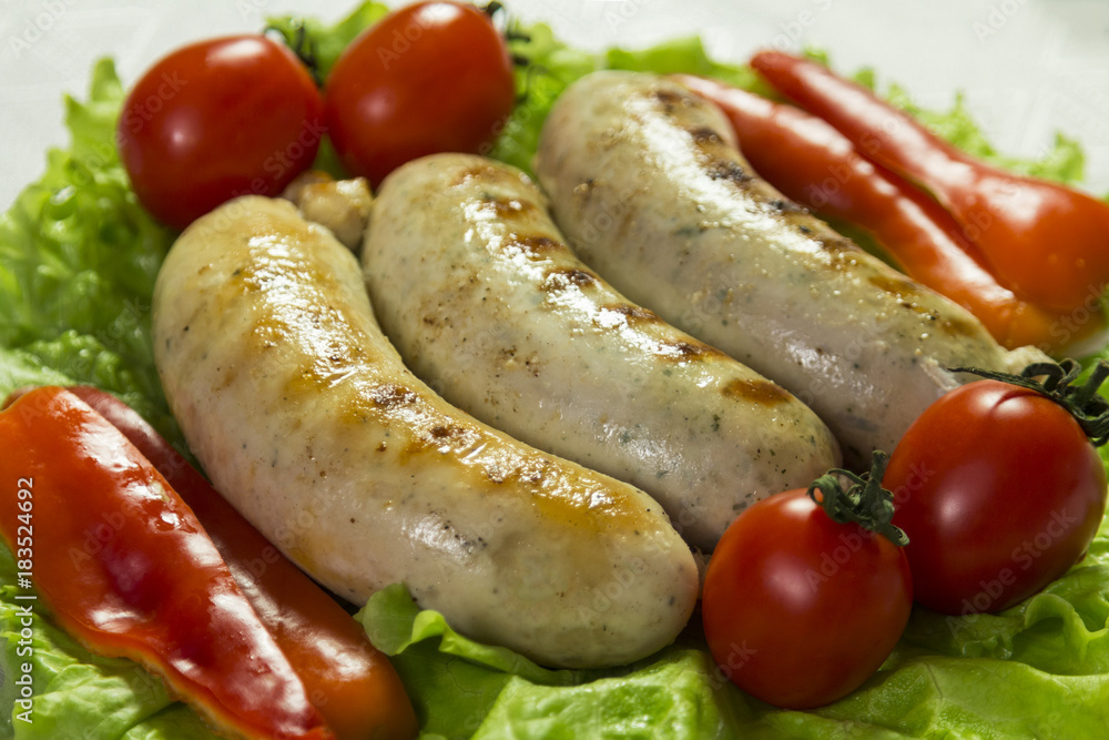 Ready-fried sausages for the grill are on the green leaves of lettuce next to tomatoes and slices of sweet pepper. Close-up. Ready-fried sausages for the grill are on the green leaves of lettuce next to tomatoes and slices of sweet pepper. Close-up.