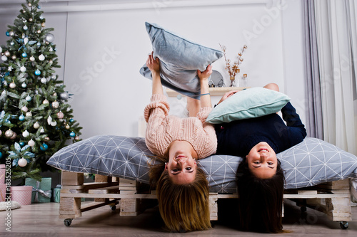 Two beautiful girls friends wear in warm sweater and leg warmers (gaiters) play with pillows on bed against new year tree with christmas decoration.