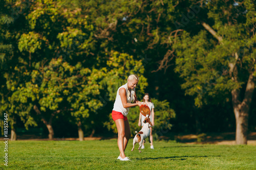 Mother and daughter throwing orange flying disk to small funny dog, which catching it on green grass. Little Jack Russel Terrier pet playing outdoors in park. Dog and women. Family resting on open air