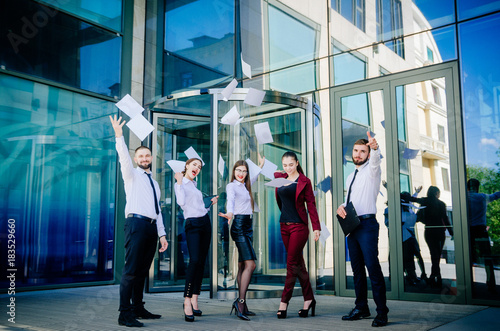 Office workers throw paper up against the background of a multi-storey glass office building. Group of young people. Five are happy about the successful completion of a profitable deal.