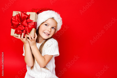 Happy smiling girl in Christmas cap holding Christmas gift in craft paper and with a red bow on a red background. Sale. Black friday. Shopping. Merry Christmas