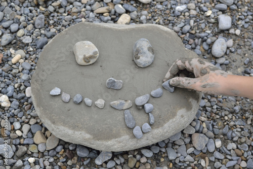 Mano de niño construyendo una cara con barro y guijarros sobre una piedra.