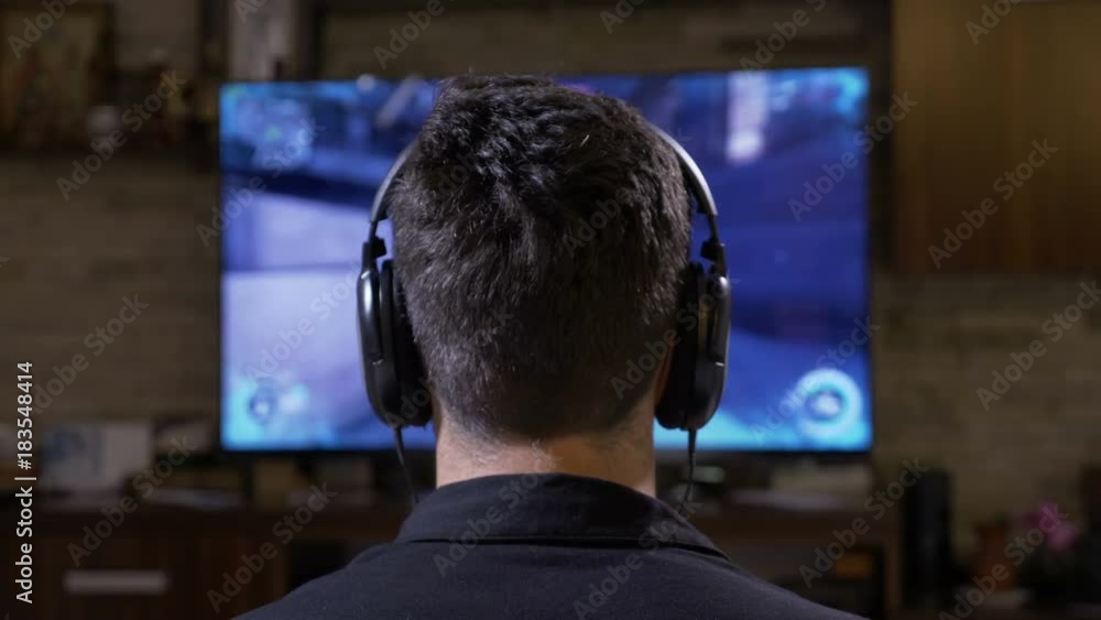Captivated young man player with big earphones relaxing at home gaming in front of digital monitor TV Captivated young man player with big earphones relaxing at home gaming in front of digital monitor TV