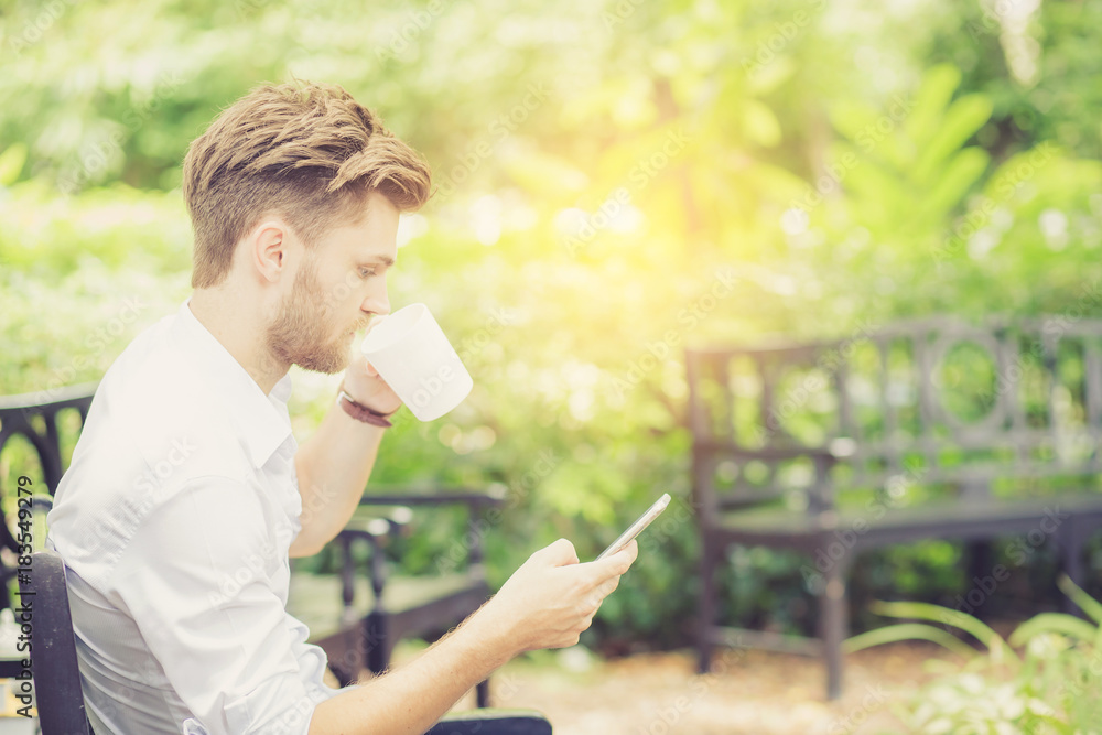 Business young handsome man drink cup of coffee using mobile phone sitting in the park, communication concept. Business young handsome man drink cup of coffee using mobile phone sitting in the park, communication concept.