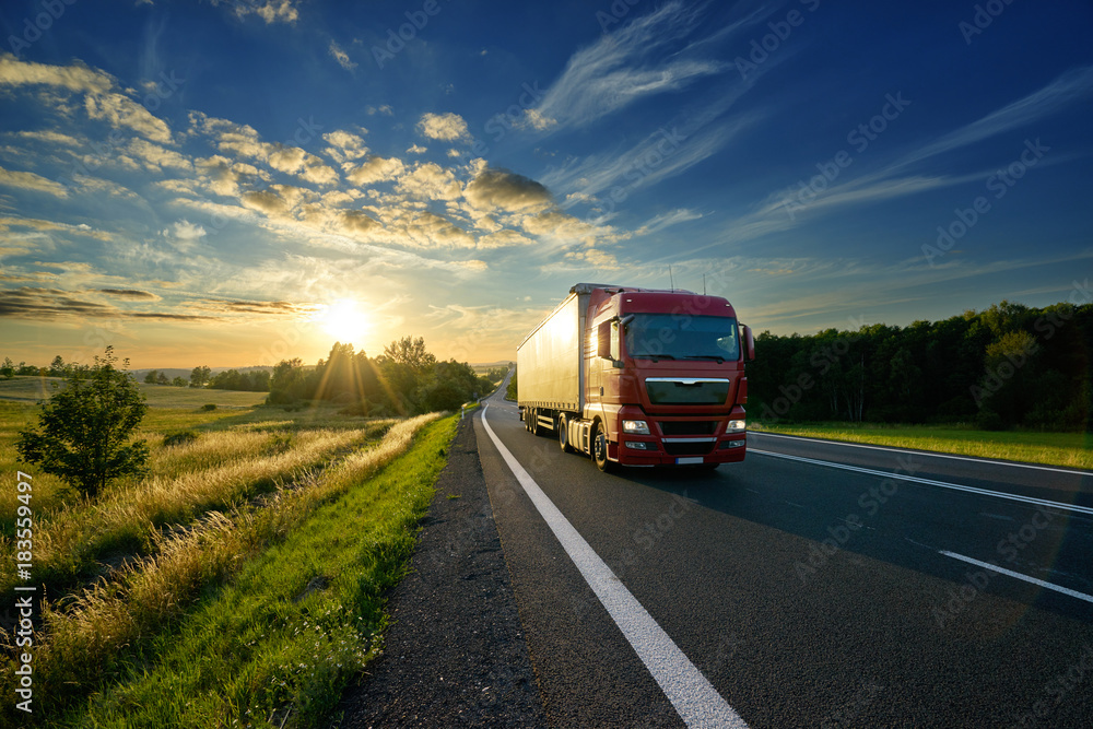 Red truck driving on the asphalt road in rural landscape at sunset