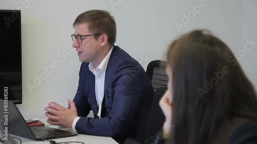 In office man and a woman are talking kindly at desk. An employee in business suit and glasses smiles and tells an interesting story to female employee at computer.