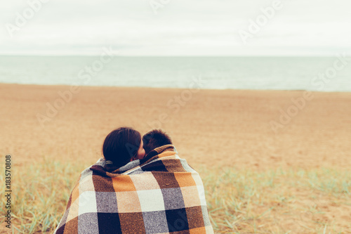 Couple sitting on the shore of natural beach wrapped up sharing a blanket on a cold winter vacation. Boyfriend and girlfriend travel lifestyle, serenity and contemplation. Vintage tonted photo. photo